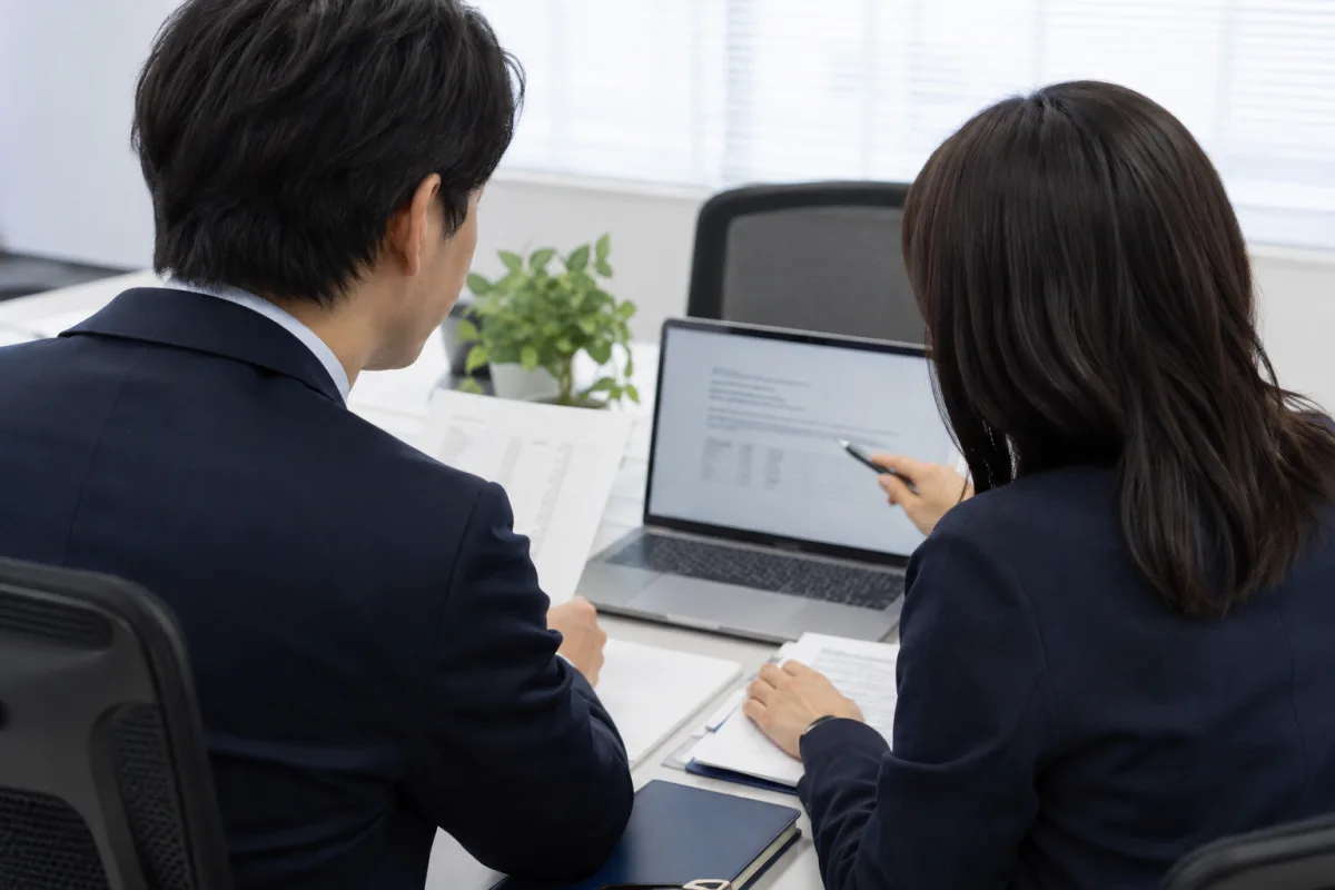 パソコンと資料を用いて企業の業務を相談する様子（AI生成のイメージ写真）